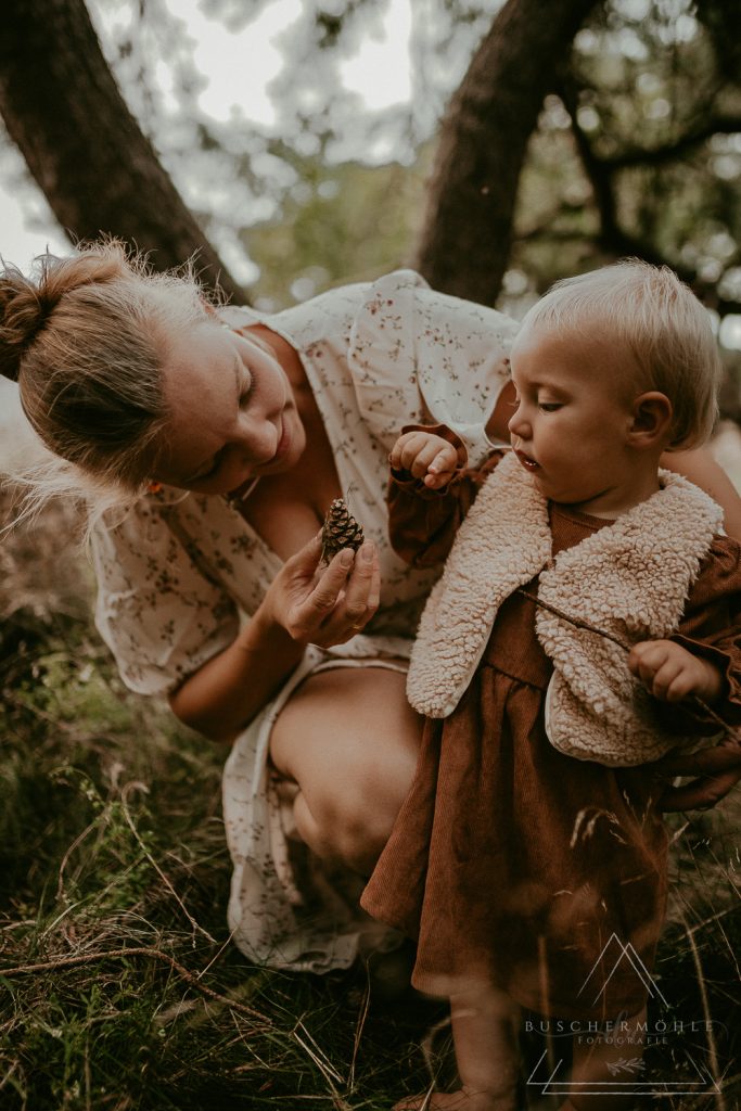 Mutter ist in der Hocke und gibt ihrem vor ihr stehenden Kleinkind einen Tannenzapfen